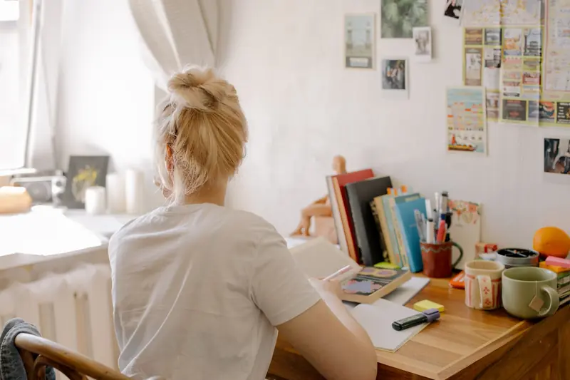 A student with blonde hair studies at a desk in a cozy dorm room setting, surrounded by books and stationery