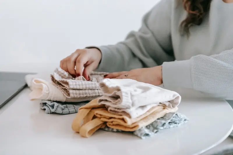 Crop faceless female in casual clothes folded colorful baby garments while sitting at white table in room on white background