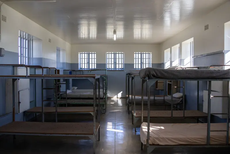 A vacant institutional dorm room with metal bunk beds and barred windows, lit by natural light