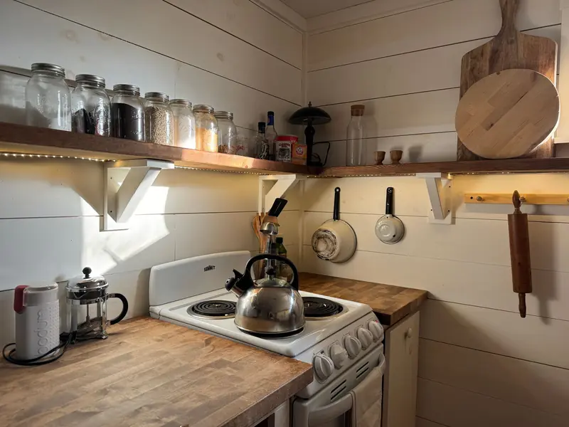 A small rustic kitchen with a wooden shelf, stove, and cooking utensils, offering a warm and cozy atmosphere